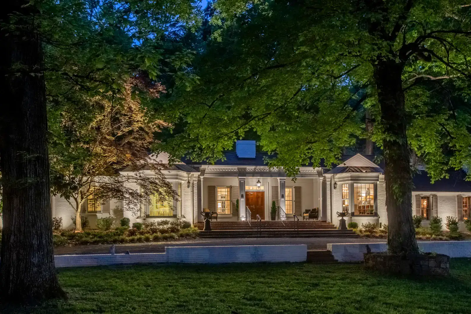 Grand columned white-brick estate with warm full-facade up-lighting, portico accent lighting, and specimen tree uplighting framed by mature hardwoods at blue hour, Belle Meade, TN
