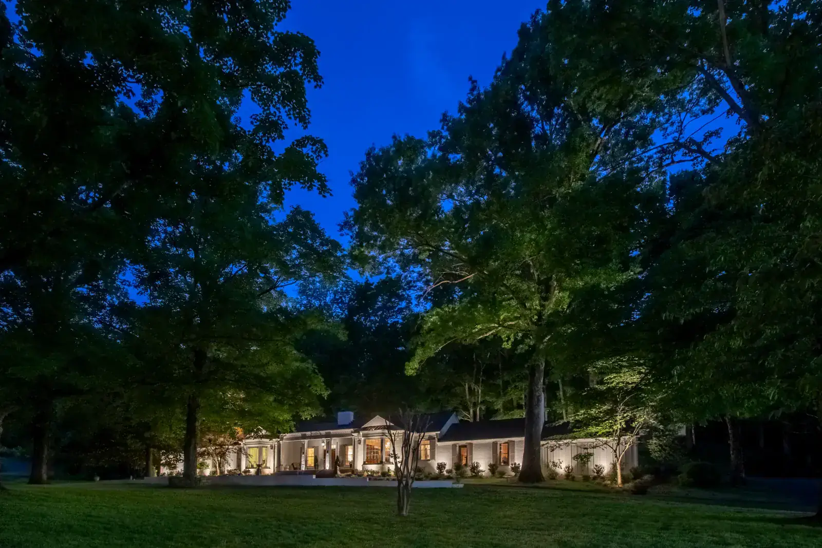 Wide-angle night view of a white-brick estate set within a mature hardwood grove with dramatic canopy tree uplighting and warm facade up-lighting at blue hour, Belle Meade, TN