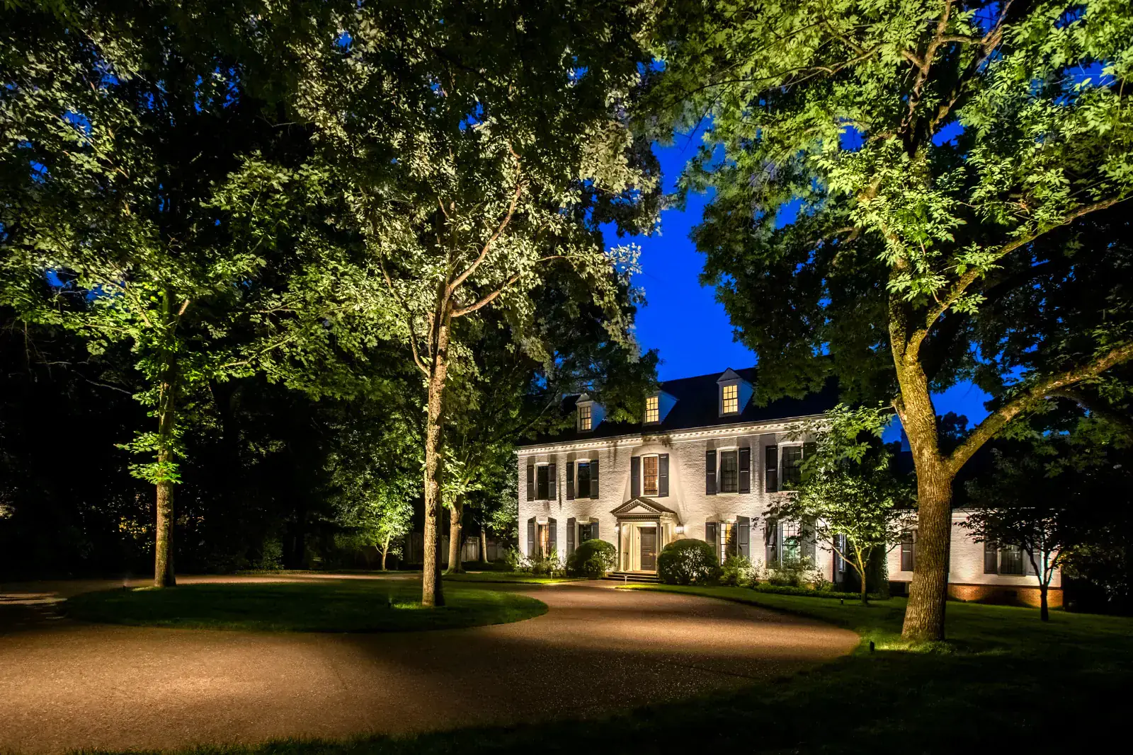 Grand white-brick Colonial estate with full canopy tree uplighting and warm facade up-lighting at blue hour, circular gravel driveway in foreground, Belle Meade, TN
