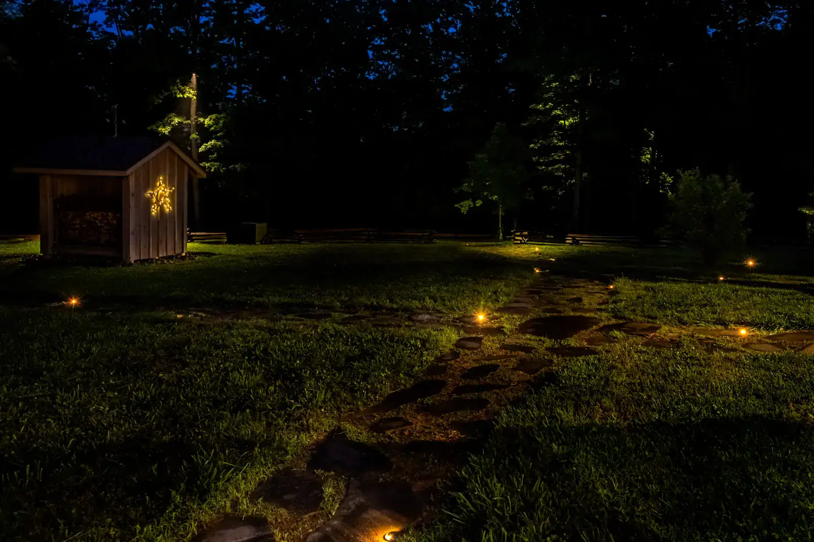 Flagstone stepping-stone path with warm in-ground path lights leading to a cedar wood shed adorned with a star and uplit trees at night, Middle Tennessee