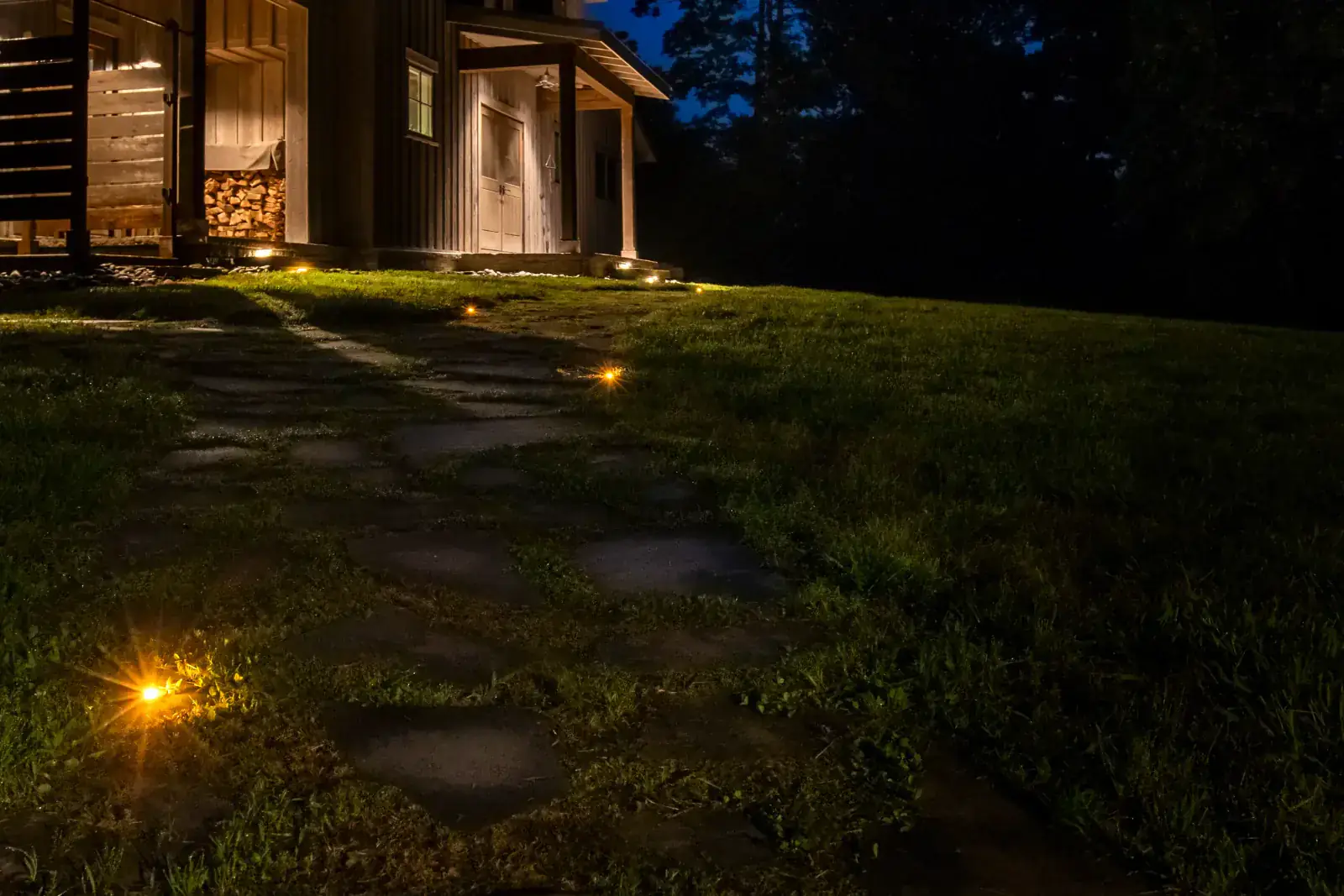 Warm LED in-ground path lights spaced along a flagstone walkway leading to the porch entry of a rustic board-and-batten cabin at blue hour, Middle Tennessee
