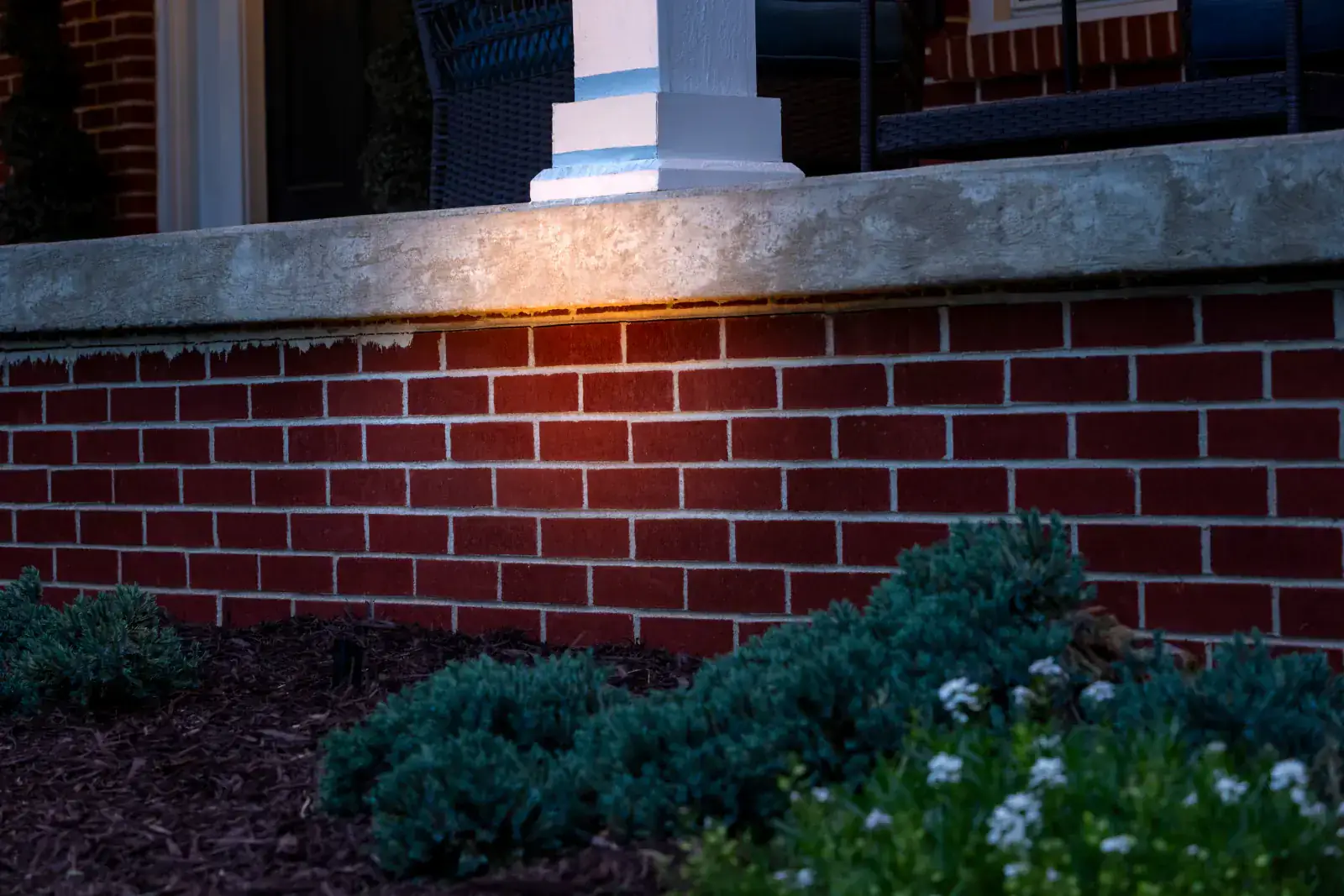 Close-up of warm LED grazing light illuminating the underside of a concrete porch beam and column base against red-brick facade with blue juniper foundation plantings at dusk, Middle Tennessee