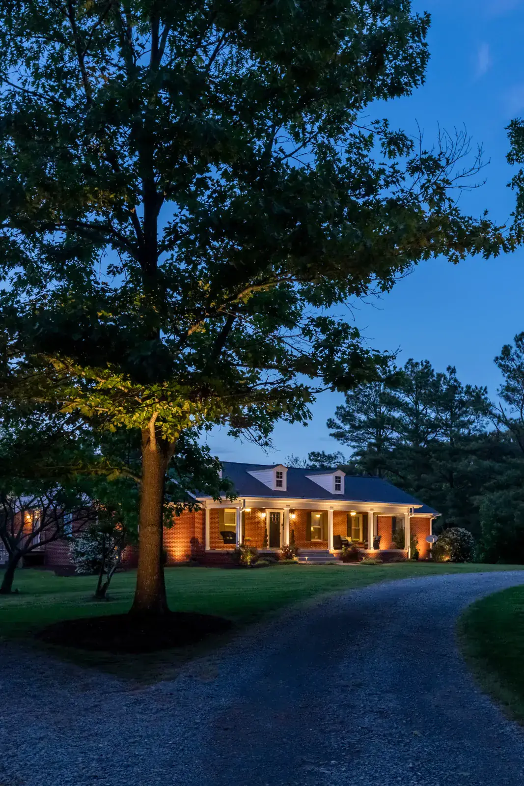 Red-brick ranch home with warm LED porch and facade up-lighting viewed through a large uplit specimen oak tree from a gravel driveway at blue hour, Middle Tennessee