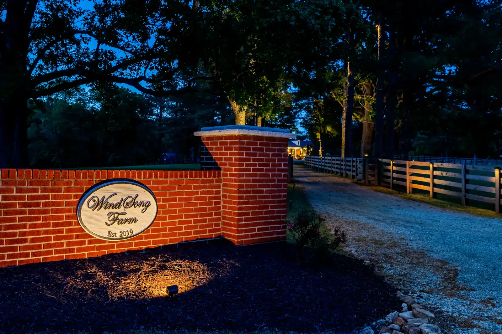 Illuminated WindSong Farm Est 2019 entrance sign on a warm LED-lit brick pillar with fence post uplighting and tree-canopy-lit gravel driveway receding to the farmhouse at blue hour, Middle Tennessee