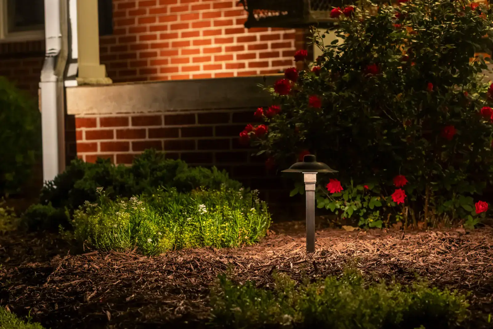 Bronze pagoda-style path light in a mulched garden bed with red roses and warm LED brick facade up-lighting at night, Middle Tennessee