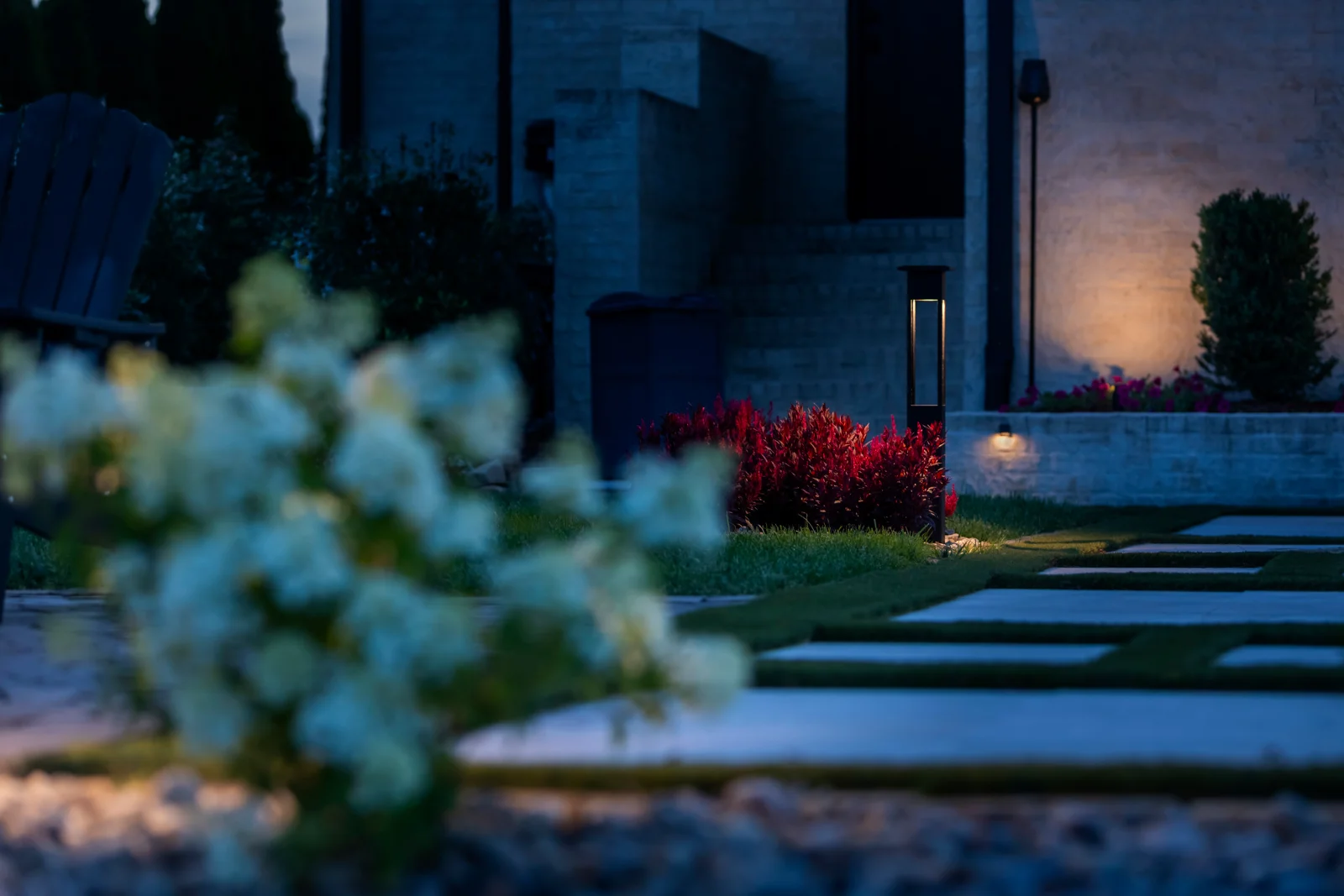 Modern dark bollard path light beside vivid red photinia shrubs with warm LED limestone facade up-lighting and stepping-stone walkway in the foreground at blue hour, Middle Tennessee