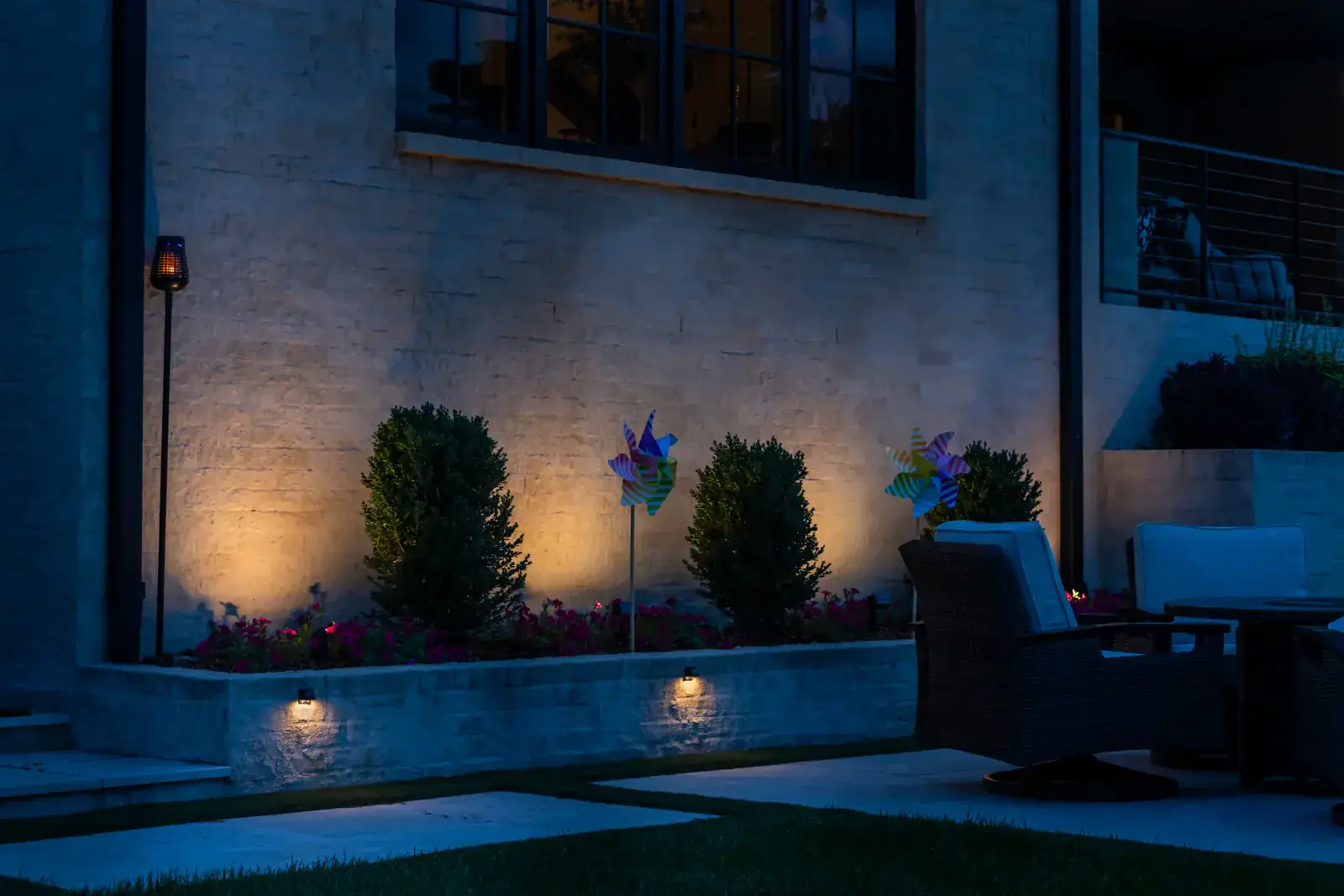 Warm LED step lights mounted in a limestone garden wall with rounded boxwood shrub up-lighting and patio outdoor seating visible at right at blue hour, Middle Tennessee