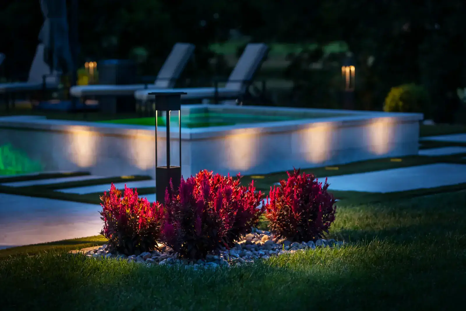 Modern dark bollard path light surrounded by vibrant red photinia shrubs with warm LED pool wall step lights and stepping-stone walkway receding into the backyard at night, Middle Tennessee