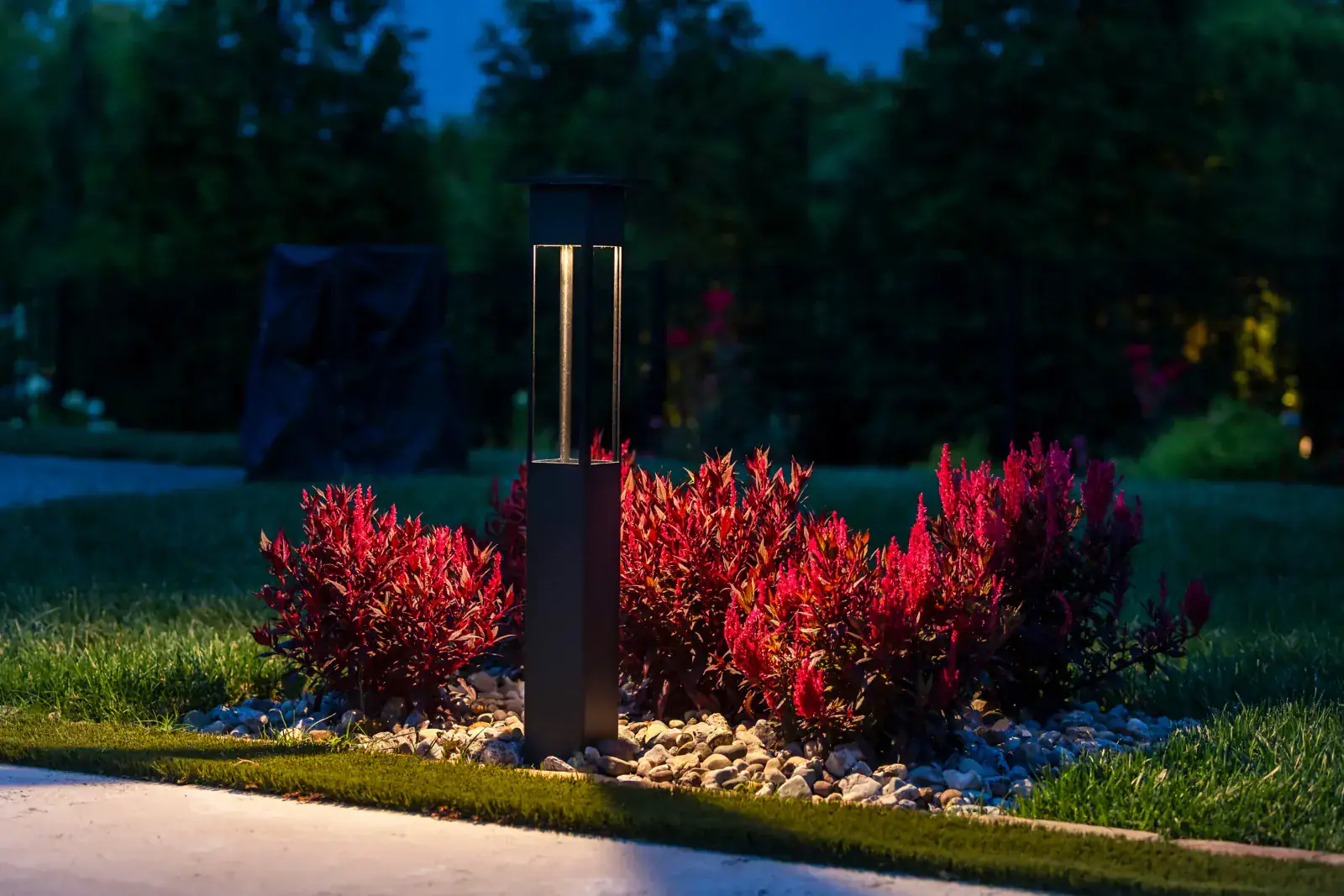 Modern dark bollard path light standing in a river-rock garden bed surrounded by vivid red photinia shrubs at blue hour, Middle Tennessee