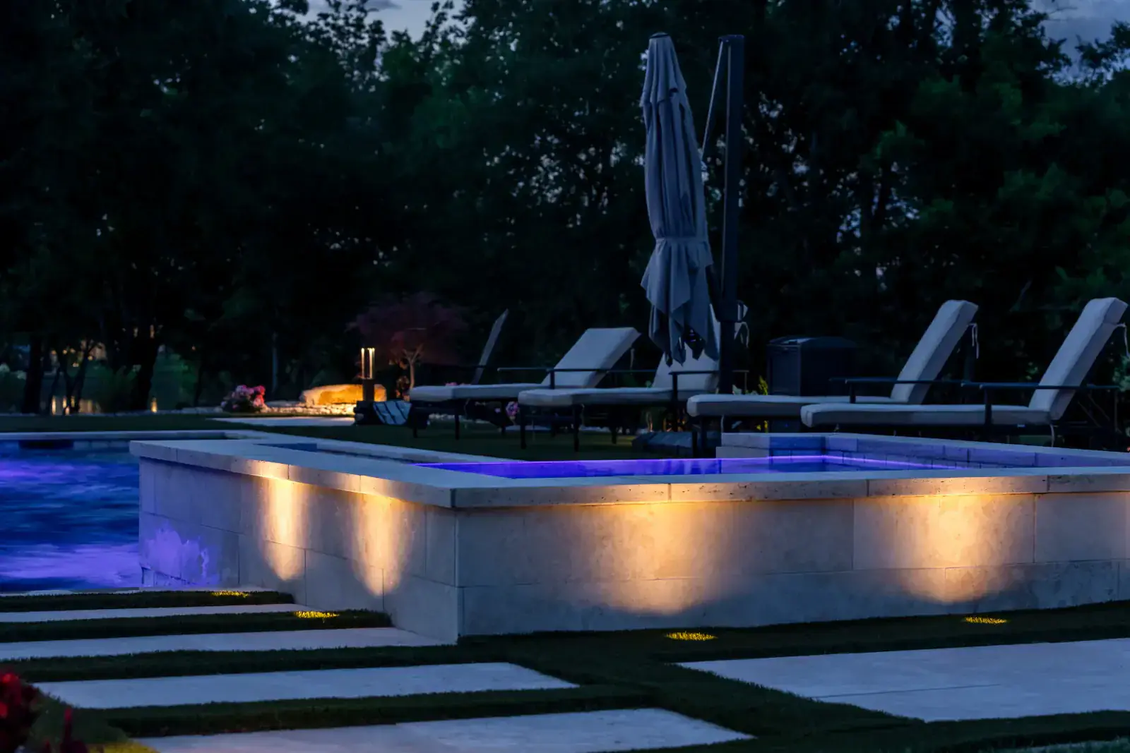 Elevated rectangular spa with warm LED up-lighting on limestone coping walls, chaise lounge chairs and pool lounge area at blue hour, Middle Tennessee