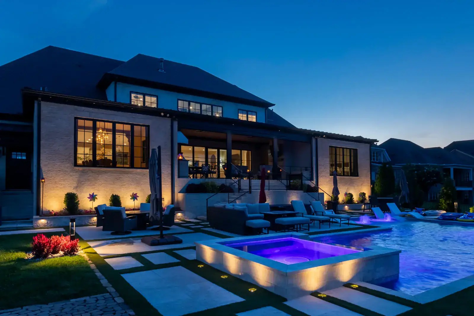 Wide-angle rear view of a modern white-brick estate with warm LED facade up-lighting, illuminated pool, purple-lit spa, pergola, and outdoor lounge area at blue hour, Middle Tennessee