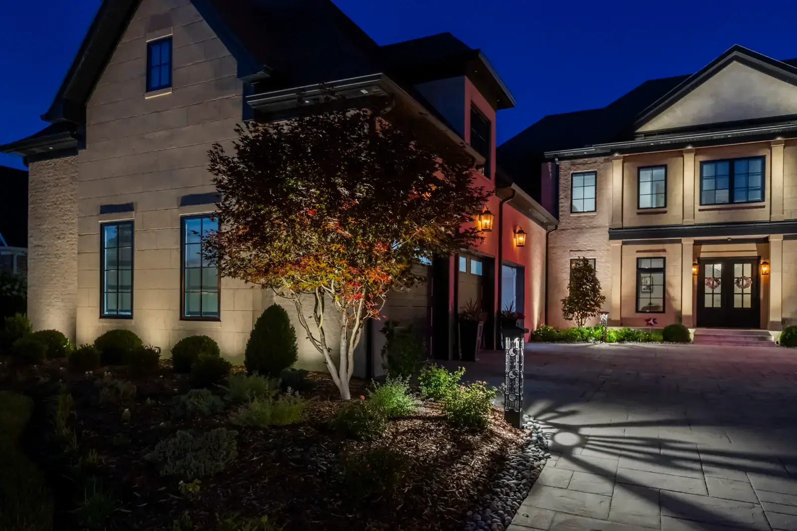 Contemporary two-tone white and stucco estate facade with warm LED facade up-lighting, dramatic Japanese maple tree uplighting, and decorative bollard path light casting shadow patterns on paver driveway at night, Middle Tennessee