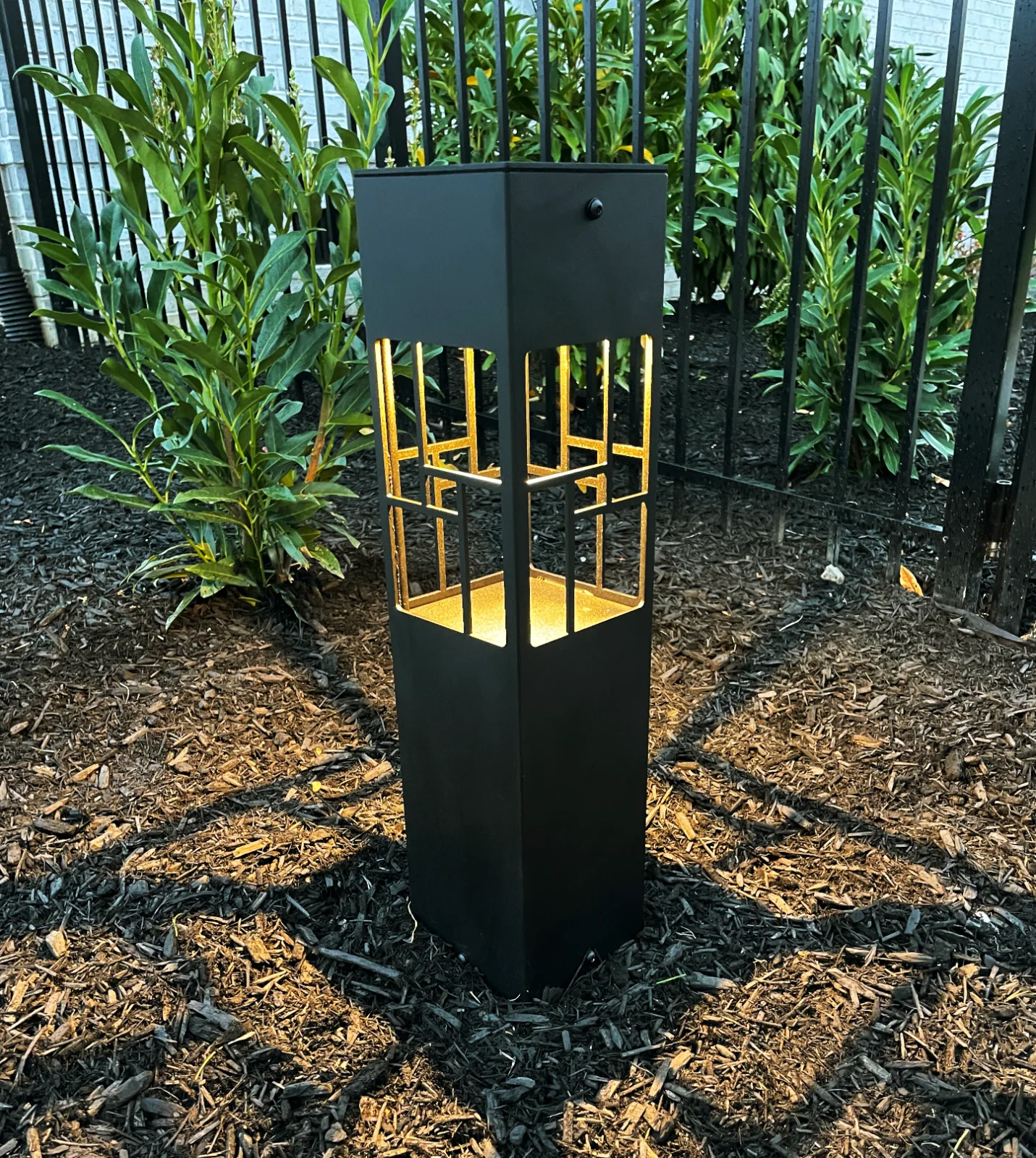 Black square bollard path light with warm amber LED glow set in a mulched landscape bed, Brentwood TN