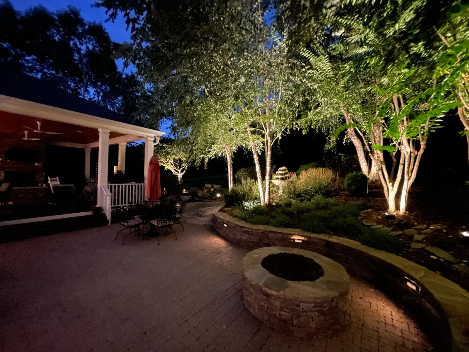 Backyard landscape lighting at blue hour in Brentwood, TN — tree uplighting, stone fire pit surround, and covered porch with warm ambient light