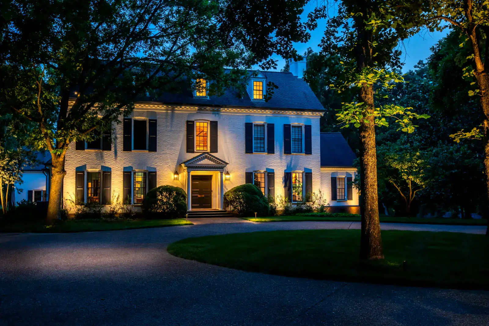Nashville estate with architectural and landscape lighting at blue hour, mature trees framing a white brick colonial facade