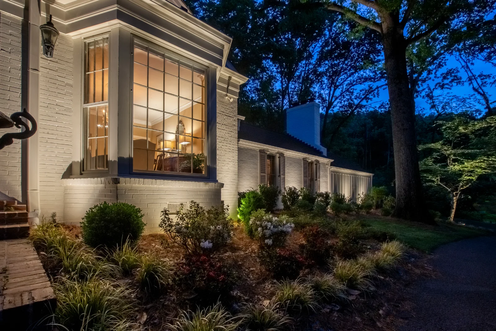 Outdoor lighting installed on a white brick home at dusk, with uplighted shrubs and landscape beds left intact